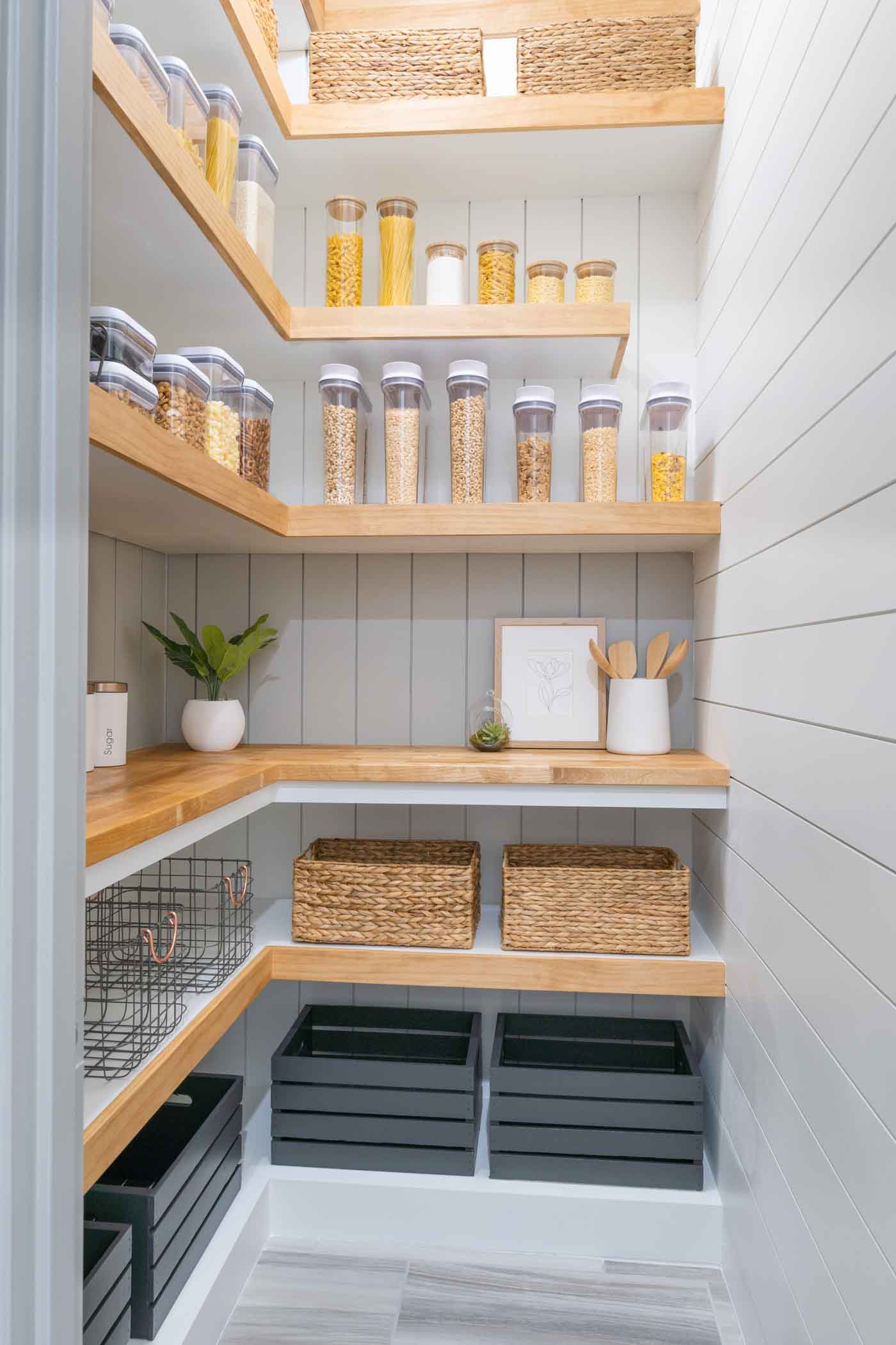 a picture of a pantry with white and gray shiplap walls, brown and white shelves, a wooden countertop, brown baskets, glass storage jars with bamboo lids, white canisters, wooden crates and metal baskets